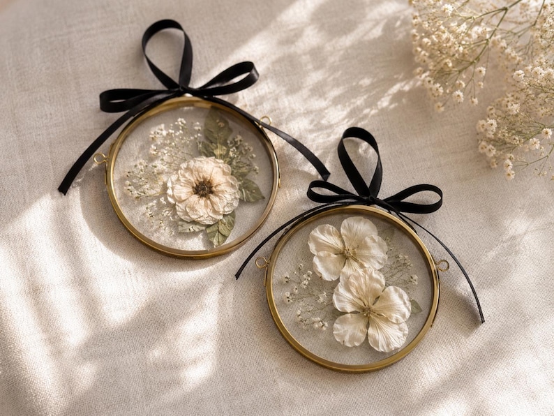 Op de afbeelding: Twee ronde messing frames met daarin geperste witte bloemen en groene bladeren achter glas. Elk frame is voorzien van een zwart satijnen lint gestrikt tot een strik, met kleine metalen ringetjes aan de zijkanten om deze decoraties op te hangen.