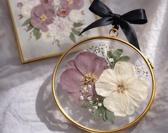 May include: A circular, gold-framed pressed flower display with a black ribbon bow. The display features a purple and cream flower arrangement with greenery. A rectangular gold-framed pressed flower art piece is in the background.