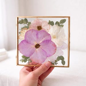Op de afbeelding: Een vierkante, goudkleurige glazen display met geperste bloemen. De compositie toont een grote, lichtpaarse bloem in het midden, omringd door witte en roze bloesems en groene bladeren. De display wordt door een hand vastgehouden.