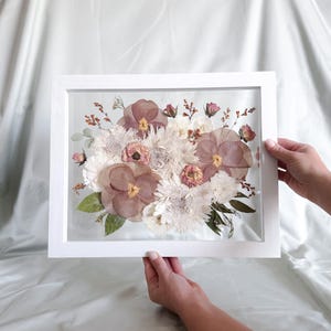 Op de afbeelding: Een rechthoekige witte lijst met een geperst bloemstuk. Het display bevat grote mauve viooltjes, witte chrysanten, kleine roze rozenknopjes en groene bladeren die tussen twee glazen platen in een zwevend ontwerp zijn geplaatst.