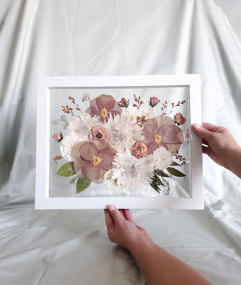 Op de afbeelding: Een rechthoekige witte lijst met een geperst bloemstuk. Het display bevat grote mauve viooltjes, witte chrysanten, kleine roze rozenknopjes en groene bladeren die tussen twee glazen platen in een zwevend ontwerp zijn geplaatst.