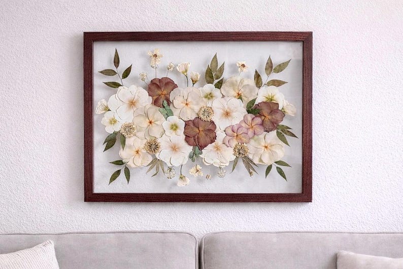 Op de afbeelding: Een rechthoekig wandobject met een verzameling geperste, gedroogde bloemen in tinten wit, cr&egrave;me en donkerbordeaux, gerangschikt met groene bladeren in een donkerbruine houten lijst met een heldere glazen voorzijde, wat een botanisch display vormt.