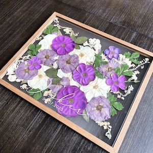 Op de afbeelding: Een rechthoekige houten lijst met een arrangement van geperste bloemen in paarse en witte tinten met groene bladeren. Op het glas staat witte cursieve tekst met de naam "Lee" en de datum "07.07.2023" over de bloemen heen gedrukt.