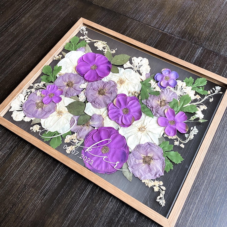 Op de afbeelding: Een rechthoekige houten lijst met een arrangement van geperste bloemen in paarse en witte tinten met groene bladeren. Op het glas staat witte cursieve tekst met de naam "Lee" en de datum "07.07.2023" over de bloemen heen gedrukt.