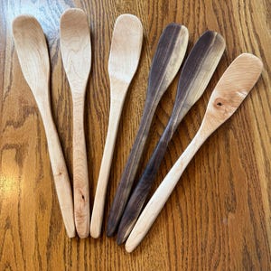 May include: Six wooden spoons of varying shades and wood grains are arranged on a wooden surface. The spoons range in color from light tan to dark brown. The spoons have long, slender handles and oval-shaped bowls. Kitchen utensils.