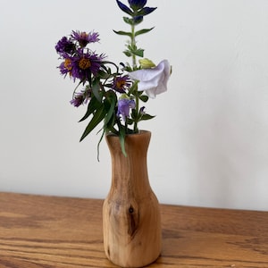 Op de afbeelding: Een kleine, handgemaakte houten vaas bevat een levendig bloemstuk. De bloemen omvatten paarse asters, een blauwe klokbloem en een witte en lavendelkleurige bloem. De vaas is gemaakt van lichtgekleurd hout en staat op een houten oppervlak.