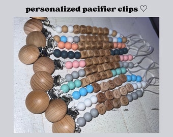 Pode incluir: Um close-up de dez prendedores de chupeta personalizados com contas de madeira e contas de silicone em várias cores. As contas de madeira são gravadas com nomes, incluindo "TUCKER", "MORGAN", "MILES", "OLIVIA", "BRYCE", "LOGAN", "MASON", "BAKER" e "TATE".