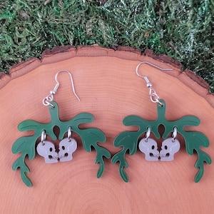 Könnte beinhalten: Ein Paar grüne Blatt-Ohrringe mit zwei grauen Totenkopf-Anhängern, die an jedem Ohrring hängen. Die Totenköpfe schauen sich an und scheinen sich zu küssen.
