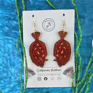 May include: Two brown glittery fish earrings with gold hooks, displayed on a white card. The card reads "California Halibut" and "Sequoia Siren". The background is blue with green glittery seaweed. A fun, ocean-themed accessory.