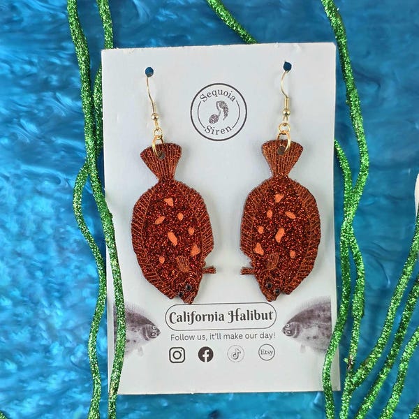 May include: A pair of brown glittery fish-shaped earrings with gold-colored hooks. The earrings are displayed on a white card with the text "California Halibut" and the brand name "Sequoia Siren". The background is blue with green glittery seaweed.