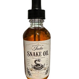 May include: A clear glass bottle with a black dropper filled with amber liquid. The label reads "Readings by Judie Team Snake Oil Spiritual Anointing Oil" with a snake illustration. The bottle contains 59 ml.