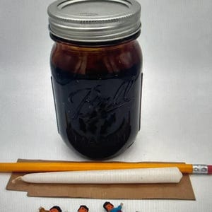 May include: A dark amber glass Ball Mason jar with a silver metal lid, a yellow pencil, a white taper candle on a brown cardboard strip, and four small handmade yarn dolls with colourful clothing arranged in a horizontal row below the items.