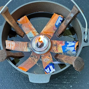 May include: A dark gray cast iron pot containing a central metal oil lamp with a lit flame. Six orange and blue Fanta soda can segments radiate outward from the center, interspersed with six large, weathered, rusted iron railroad spikes.