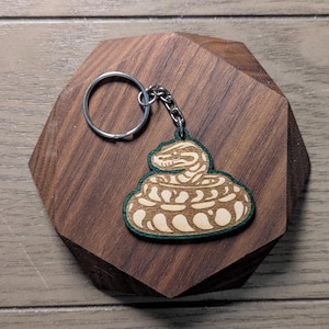 May include: A wooden keyring with a coiled snake design in light brown and green. The keyring is attached to a silver metal ring and chain. The snake design is displayed on a wooden, geometric stand.