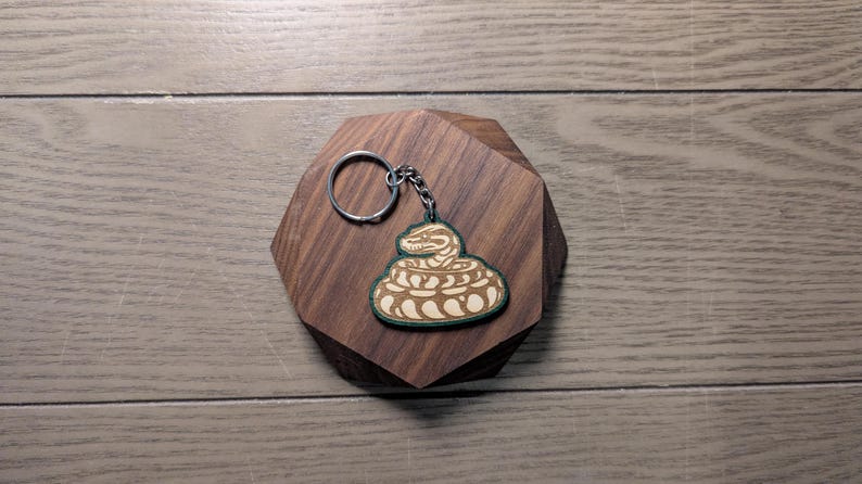 Op de afbeelding: Een houten sleutelhanger met een opgerold slangontwerp in lichtbruin en groen. De sleutelhanger is bevestigd aan een zilveren metalen ring en ketting. Het slangontwerp wordt weergegeven op een houten, geometrische standaard.