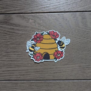 Könnte beinhalten: Ein Aufkleber mit einem gelben Bienenstock mit schwarzem Eingang, umgeben von roten Blumen und zwei Bienen. Die Bienen haben schwarz-gelbe Streifen und weiße Flügel. Der Aufkleber befindet sich auf einer Holzoberfläche.