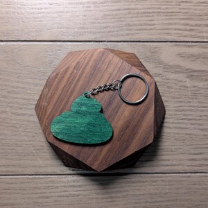 Op de afbeelding: Een groene houten sleutelhanger in de vorm van een wolk rust op een geometrisch houten blok. De sleutelhanger heeft een zilveren ketting en ring. Het blok is gemaakt van donkerbruin hout en ligt op een lichtbruin houten oppervlak.