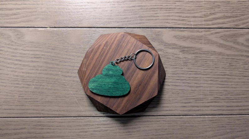 Op de afbeelding: Een groene houten sleutelhanger in de vorm van een wolk rust op een geometrisch houten blok. De sleutelhanger heeft een zilveren ketting en ring. Het blok is gemaakt van donkerbruin hout en ligt op een lichtbruin houten oppervlak.
