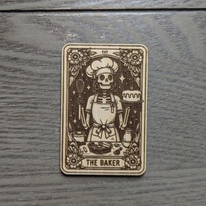 May include: A wooden card with a skeleton baker wearing a chef's hat and holding a whisk and a cake. The card is decorated with flowers and stars. The text "EST" and "THE BAKER" are printed on the card.