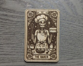 May include: A wooden card with a skeleton baker wearing a chef's hat and holding a whisk and a cake. The card is decorated with flowers and stars. The text "EST" and "THE BAKER" are printed on the card.