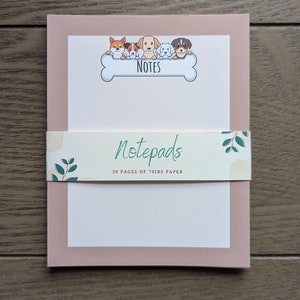 May include: A pink notepad with a white interior and a border around the edges. The notepad has a cartoon image of five dogs peeking over a bone with the word "NOTES" written on it. The notepad has a sticker on the front that says "Notepads 30 pages of 70lbs paper".