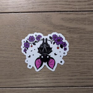 May include: A white sticker featuring a black bat hanging upside down from a branch with purple flowers and red berries. The bat has pink ears and is surrounded by small stars and bats.