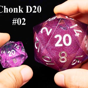 May include: Two purple D20 dice with glitter. The larger die has the number 20 on one side and the smaller die has the number 20 on one side. The larger die is labeled "Chonk D20 #02".