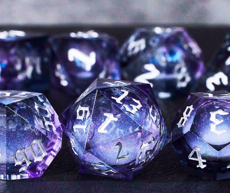 Puede incluir: Un conjunto de ocho dados de color morado y plateado con un patr&oacute;n de galaxia. Los dados est&aacute;n hechos de pl&aacute;stico transparente con un acabado brillante. Los n&uacute;meros en los dados son plateados y est&aacute;n delineados en negro.