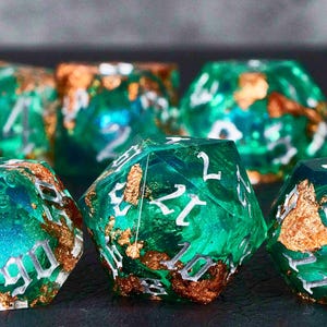 May include: A set of green and gold dice with white numbers. The dice are made of resin and have a speckled appearance. The dice are perfect for tabletop gaming.
