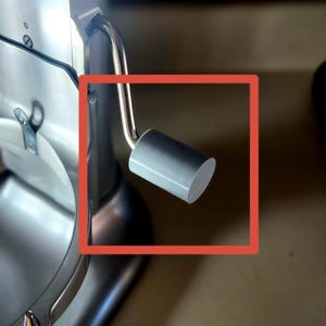 May include: A close-up of a silver stand mixer with a gray handle. The mixer has a rounded base and a curved arm. The handle is cylindrical and gray. The mixer is on a light brown surface.