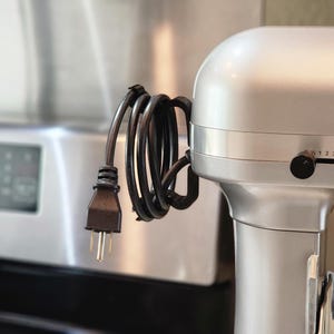 May include: A silver stand mixer with a black power cord wrapped around it. The cord has a three-prong plug. The mixer has a dial with numbers 1, 2, and 3. The background is a blurred kitchen scene.
