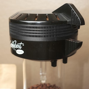Könnte beinhalten: Ein schwarzer Kaffeeröster mit einer transparenten Glaskammer, die mit gerösteten Kaffeebohnen gefüllt ist. Der Röster hat den Text "Fresh Roast SR800" an der Seite. Die Oberseite hat Lüftungsschlitze und einen Griff.