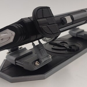 May include: A black and gray 3D printed replica of a futuristic weapon, displayed on a stand with a circular design. The weapon has a sharp, pointed tip and a textured surface.