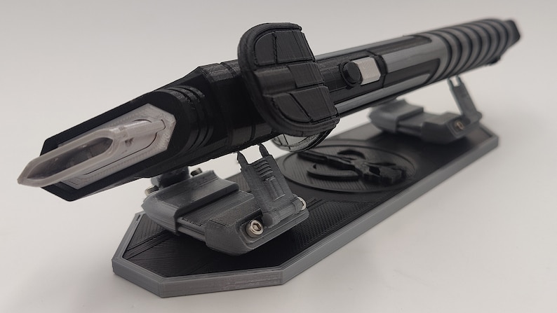 May include: A black and gray 3D printed replica of a futuristic weapon, displayed on a stand with a circular design. The weapon has a sharp, pointed tip and a textured surface.
