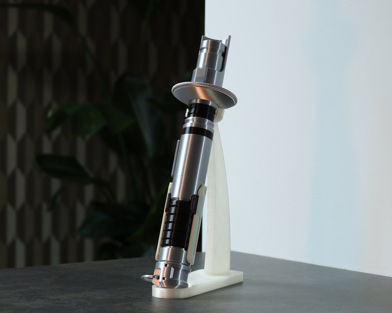 May include: A silver lightsaber replica with a black handle and a white stand. The lightsaber is on a dark gray surface.