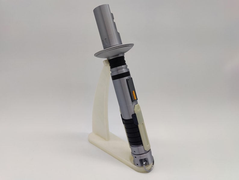 May include: A silver lightsaber with black and white accents on a white stand. The lightsaber has a black handle and a silver blade. The stand is made of plastic and has a triangular shape.