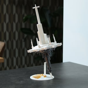 May include: A white 3D printed model of a futuristic city with a tall spire, floating on a dark grey base. The model is on a white circular stand with a gold accent.