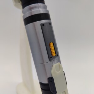 May include: A silver and black lightsaber with a yellow button on the side. The lightsaber is made of metal and has a textured surface.