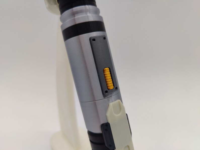 May include: A silver and black lightsaber with a yellow button on the side. The lightsaber is made of metal and has a textured surface.