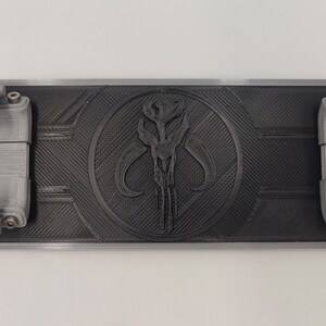 May include: A black 3D printed wall mount with a Mandalorian symbol in the center. The mount has two gray, 3D printed arms on either side.