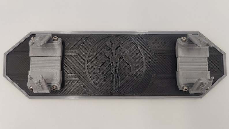 May include: A black 3D printed wall mount with a Mandalorian symbol in the center. The mount has two gray, 3D printed arms on either side.
