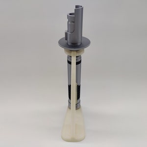 May include: A white plastic stand with a gray lightsaber on top. The lightsaber is in a vertical position and has a black and gray design.
