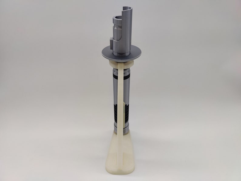 May include: A white plastic stand with a gray lightsaber on top. The lightsaber is in a vertical position and has a black and gray design.