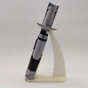 May include: A silver lightsaber prop with a black and white handle, displayed on a white plastic stand.