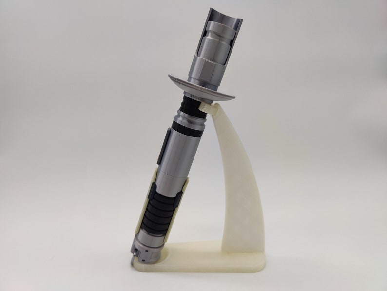 May include: A silver lightsaber prop with a black and white handle, displayed on a white plastic stand.