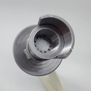 May include: A close-up of a silver metal 3D printed object with a circular opening and a series of small holes. The object has a rounded, curved top.