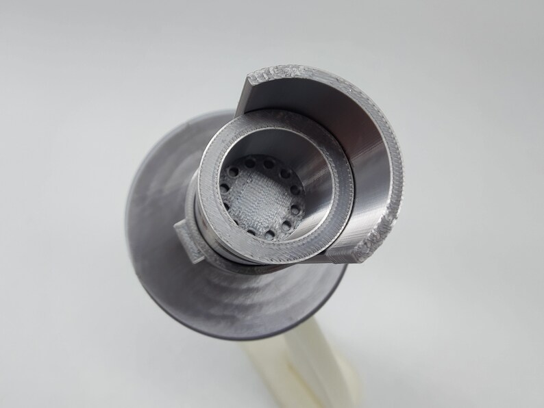 May include: A close-up of a silver metal 3D printed object with a circular opening and a series of small holes. The object has a rounded, curved top.