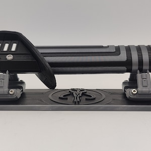 May include: A black and silver lightsaber with a Mandalorian symbol on the stand. The lightsaber is on a display stand with a black and silver finish.