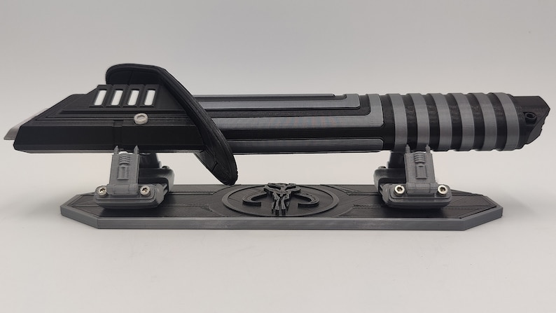 May include: A black and silver lightsaber with a Mandalorian symbol on the stand. The lightsaber is on a display stand with a black and silver finish.