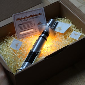 May include: A silver lightsaber prop in a cardboard box with shredded paper. The box is open and the lightsaber is lit up with a warm yellow glow. The box also contains a thank you card and a small business card.